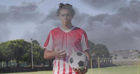 Young soccer player standing in rain holding ball on stormy outdoor football field practice