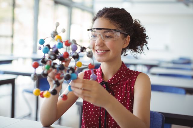 Young schoolgirl wearing safety goggles holding a colorful molecular model in a science lab. Ideal for educational content, STEM promotion, classroom activities, and science-related materials.