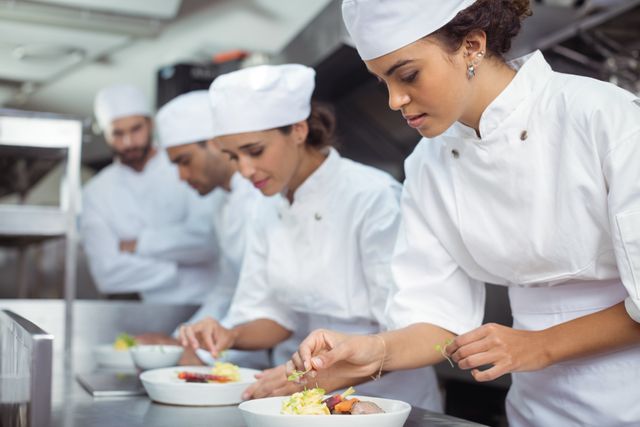 Female chef garnishing a dish in a professional kitchen with other chefs working in the background. Ideal for use in articles or advertisements related to culinary arts, restaurant industry, teamwork in kitchens, and professional cooking. Suitable for illustrating concepts of food preparation, gourmet cuisine, and culinary skills.