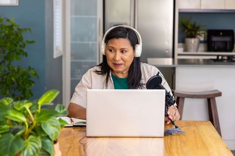 Mature woman recording podcast from home kitchen with laptop microphone and headphones