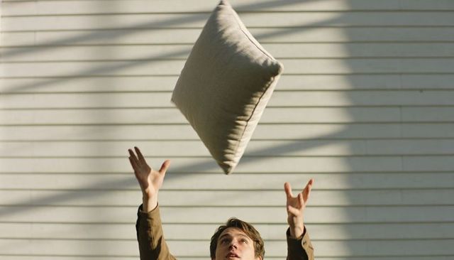 Young man tossing square pillow on sunlit porch, reaching up to diagonal siding shadows