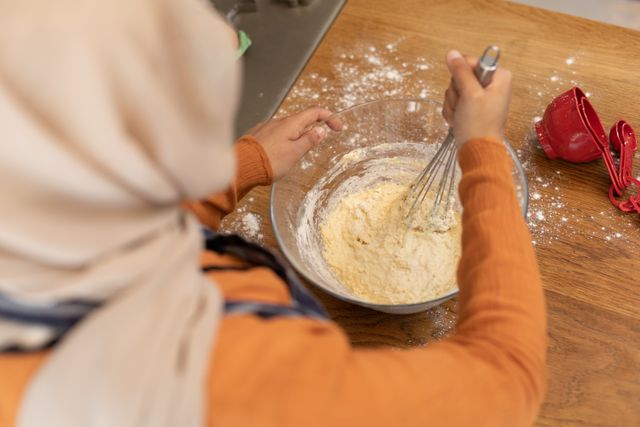 Biracial woman in hijab mixing dough in a bowl in a kitchen. Ideal for use in articles or advertisements about inclusivity, domestic life, cooking, and cultural diversity. Can also be used in content promoting home baking, recipes, and family activities.