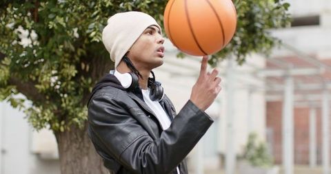 Young man spinning basketball on finger in urban courtyard wearing beanie and headphones