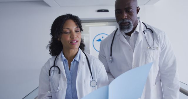 Two doctors are seen reviewing a patient's chart while walking down a hospital corridor. Both are wearing white lab coats and stethoscopes. The woman doctor is holding the chart and pointing out details while conversing with her male colleague. This can be used for healthcare, medical teamwork, and professional collaboration content.