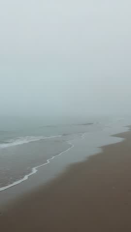 Vertical foggy shoreline video with gentle waves lapping sandy beach and thin foam lines