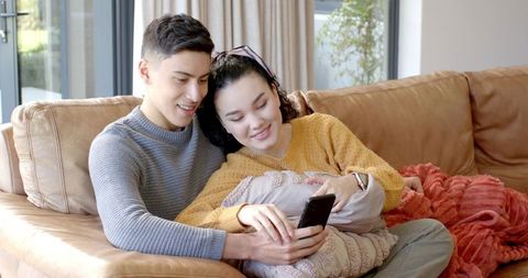 Asian Couple Relaxing on Sofa Sharing Smartphone in Bright Living Room