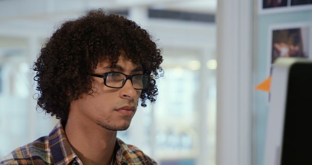 Young man with curly hair wearing glasses working intently on computer. This image can be used to represent professionals, technology usage, office work, focus, academic tasks, and workplace environments.