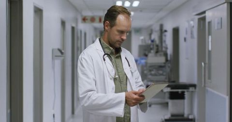 Male Doctor Reviewing Clipboard with Stethoscope in Hospital Corridor