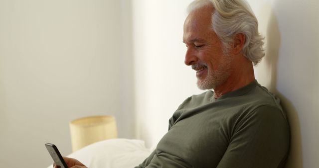 Smiling senior man dressed in casual clothing relaxing on bed while using smartphone. White-haired man enjoying leisure time with technology. Ideal for themes related to senior lifestyle, modern technology, happiness, and relaxation at home.