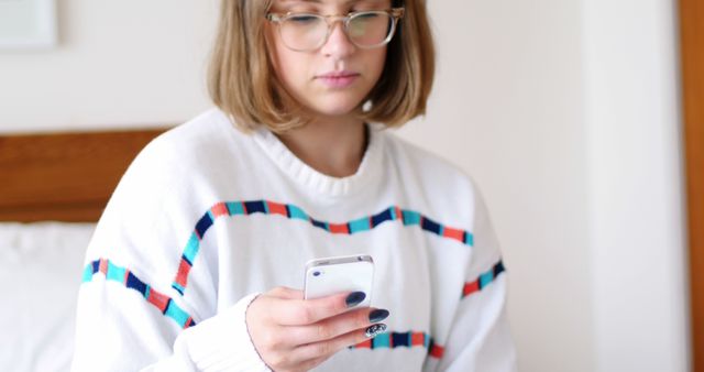 Woman sitting on a bed, using a smartphone while wearing glasses, suggests focus and engagement with technology in a relaxed home environment. Great for illustrating casual digital use, home relaxation, or modern communication.