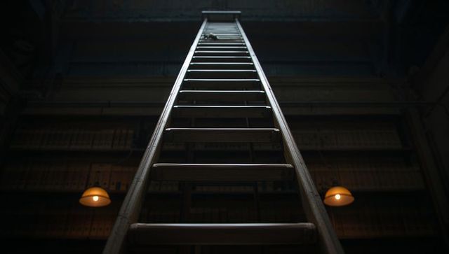 Rising tall wooden ladder reaching hatch in moody archive with amber pendant lighting