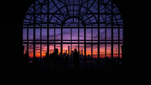 Dramatic sunset skyline framed by grand arched terminal window with silhouetted commuters