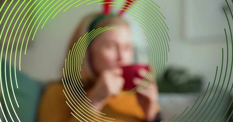 Mature woman sipping red mug wearing antler headband on couch with green circular overlay