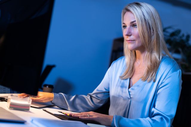 Caucasian businesswoman with blond hair working on computer in office at night. Ideal for illustrating concepts of dedication, productivity, and corporate work environments. Suitable for articles on work-life balance, business technology, and professional commitment.