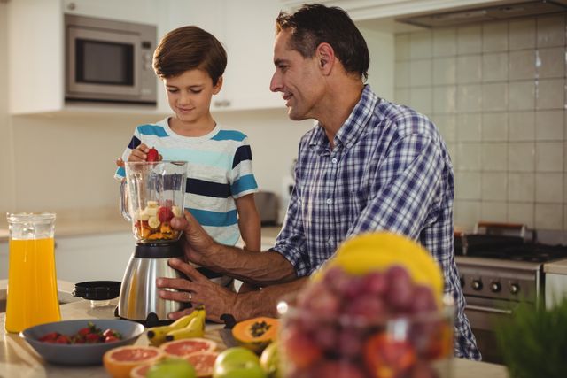 Father and son are preparing a smoothie together in a modern kitchen. The father is assisting his son in adding fruits to a blender. The kitchen counter is filled with fresh fruits and a glass of orange juice. This image can be used for promoting healthy family lifestyles, parenting tips, cooking classes, and nutritional guides.