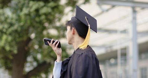Asian graduate speaking into smartphone on campus wearing cap and gown with yellow tassel