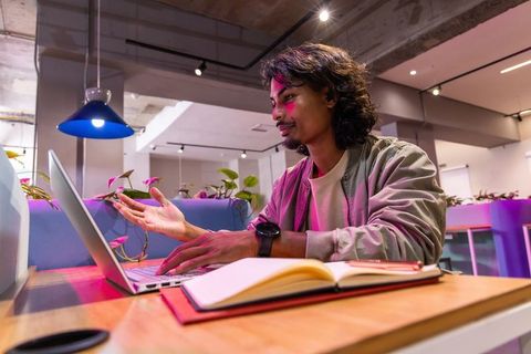 Indian man in twenties typing and gesturing during virtual meeting in colorful coworking