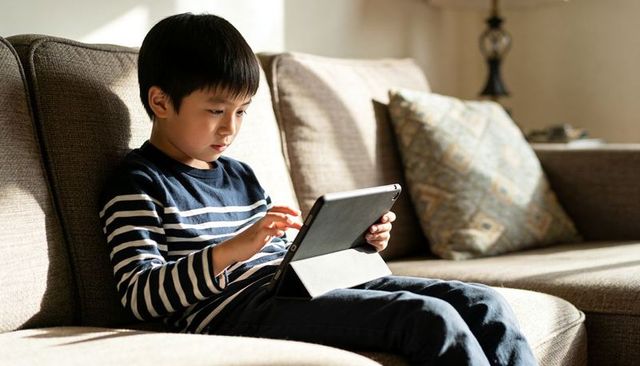 Young boy using tablet on cozy sofa with warm sunlight, learning and relaxing at home