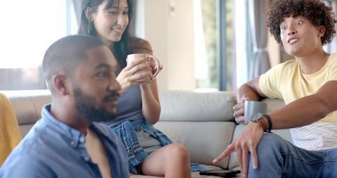 Diverse friends chatting on sunlit sofa with coffee and smartphone, relaxed home hangout