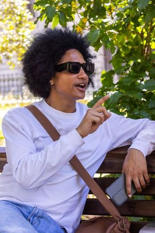 African american man sitting on park bench pointing while holding smartphone wearing sunglasses