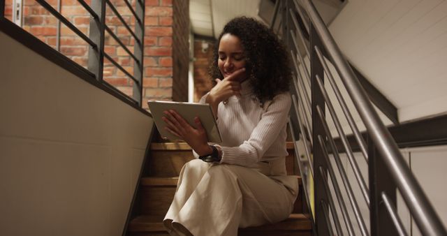 Young woman, sitting on stairs, working on her tablet in a modern office space. She appears concentrated and engaged with her work. Great for use in business, technology, and modern workplace topics. Relevant for presentations on remote work, freelance opportunities, or digital work environments.