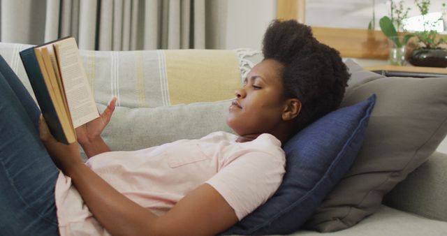 Woman is relaxing on a couch in a cozy living room, engaged in reading a book. She looks at ease and comfortable. This image can be used in articles or advertisements about relaxation, home comfort, reading habits, leisure activities, or lifestyle content related to young women and self-care.