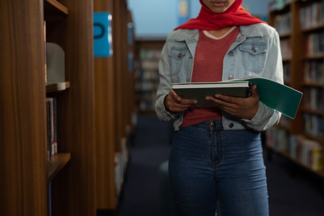 Front view mid section of an Asian female student wearing a red hijab and jeans studying in a library, holding a book in hands.