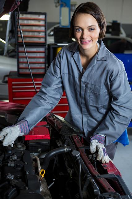 Female auto mechanic wearing gray uniform and gloves, smiling while repairing engine in a garage. Ideal for use in articles or advertisements about gender diversity in industries, automotive repair services, and professional women in male-dominated fields.