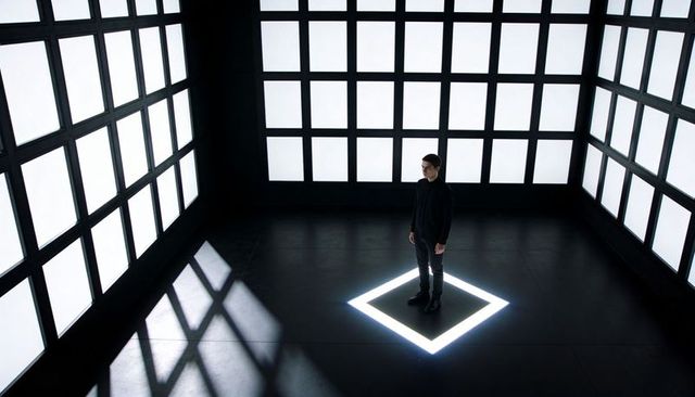 Solitary man standing in illuminated floor square amid grid light panels minimal monochrome