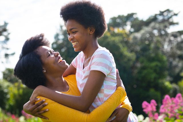 This image captures a joyful moment between an African American mother and her teenage daughter, smiling and enjoying each other's company outdoors on a sunny day. Ideal for use in family-oriented advertisements, parenting blogs, lifestyle articles, and promotional materials emphasizing family bonding, happiness, and togetherness.