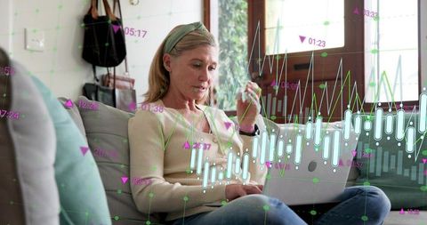 Mature woman analyzing stock charts on laptop at home wearing smartwatch, casual sweater