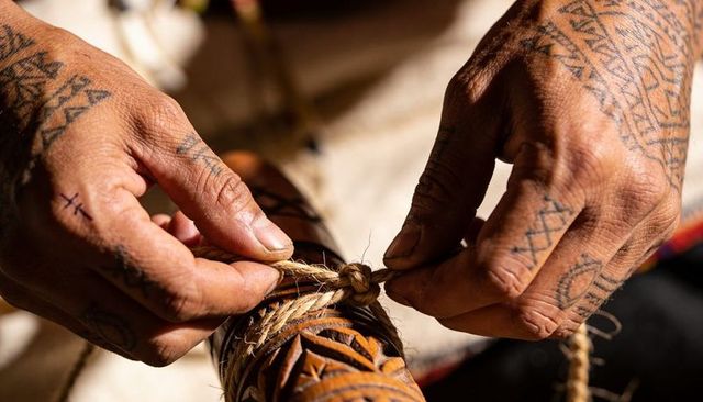 Tattooed hands tying twine around carved wooden cylinder crafting traditional knot