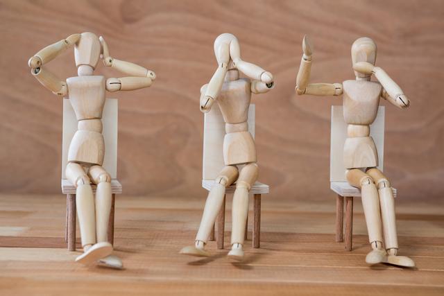 Three wooden mannequins are sitting on chairs, each representing one of the three wise monkeys: see no evil, hear no evil, and speak no evil. This conceptual image can be used in articles or presentations about communication, wisdom, philosophy, or human behavior. It is also suitable for illustrating concepts related to non-verbal communication and body language.