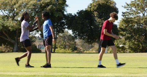 Young friends high-fiving during jog in park while teammate walks on sunny field