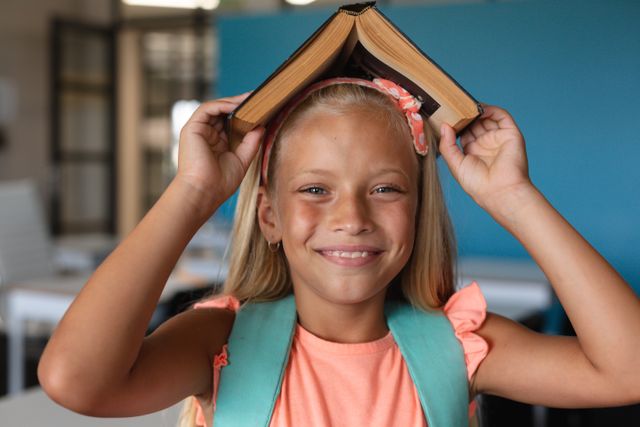 Portrait of smiling caucasian elementary schoolgirl holding book on head while sitting in class. unaltered, childhood, education, playful and school concept.