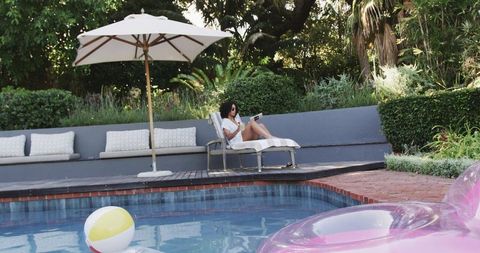 African american woman relaxing on lounge chair reading by pool with pink float