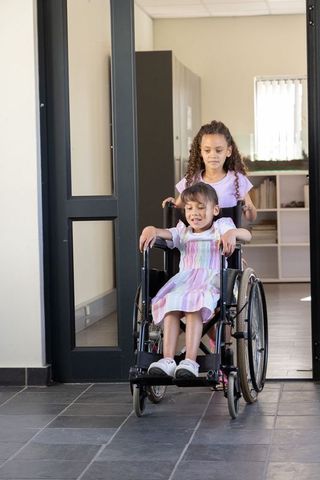 School peers pushing wheelchair with smiling child in pastel dress promoting inclusion