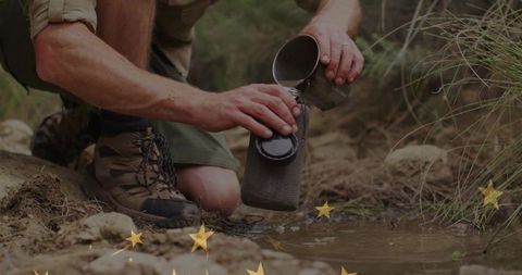 Kneeling hiker refilling canteen at creek edge pouring water from metal cup outdoors
