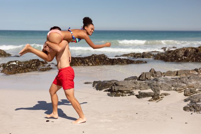 Young man carrying woman on shoulders while enjoying a sunny day at the beach. Perfect for use in advertisements for travel agencies, summer vacation promotions, lifestyle blogs, and social media posts celebrating love and fun.