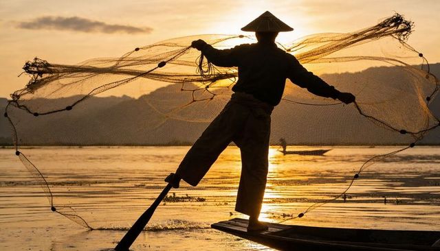 Silhouette fisherman casting circular net at golden sunset, balancing on wooden boat with reflection