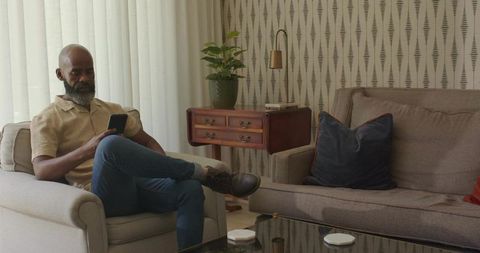 Middle-aged man sitting on armchair using smartphone in modern living room with calm decor