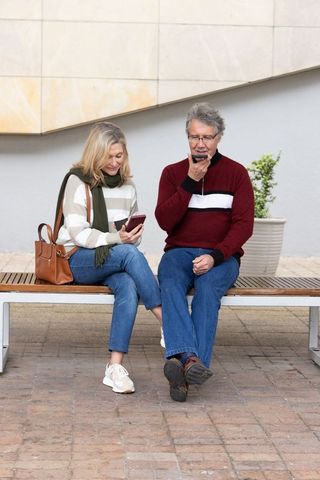 Senior couple sitting on bench using smartphones and sharing casual moment in urban plaza