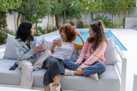 Diverse friends chatting and sipping coffee on outdoor sofa by poolside backyard terrace