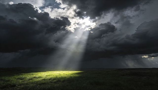 Sunbeams Piercing Dark Storm Clouds Revealing Lush Sunlit Grass Patch on Open Plain
