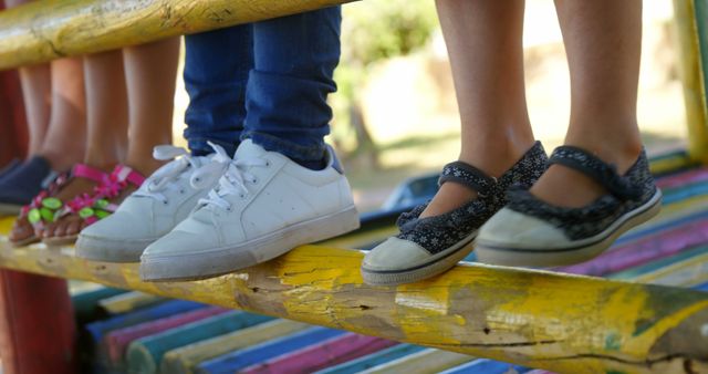 Feet of diverse children wearing different styles of shoes are perched on a colorful wooden beam, with copy space. It captures a moment of casual playfulness and the simplicity of childhood.
