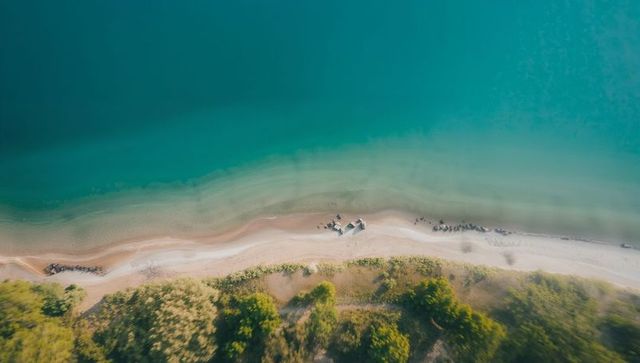 Turquoise aerial shoreline with sandy beach and dunes, clustered rocks and gentle groynes