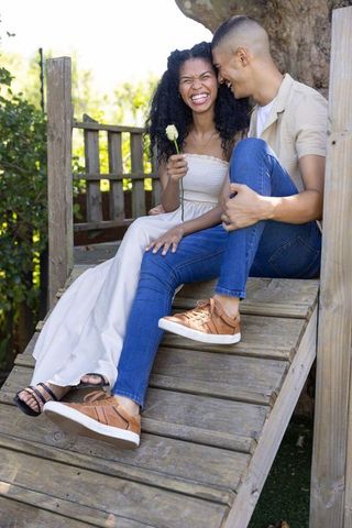 Laughing couple sitting on backyard wooden ramp sharing white rose and playful affection