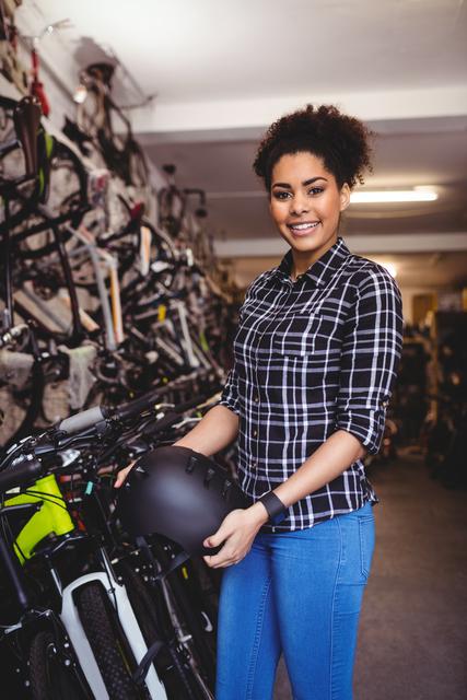Young female mechanic holding a bicycle helmet in a workshop filled with bicycles. She is smiling and wearing a plaid shirt and jeans. Ideal for use in articles about women in trades, bicycle maintenance, cycling safety, and professional workshops.