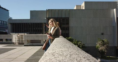 Young woman leaning on concrete balustrade holding coffee and smartphone on urban plaza