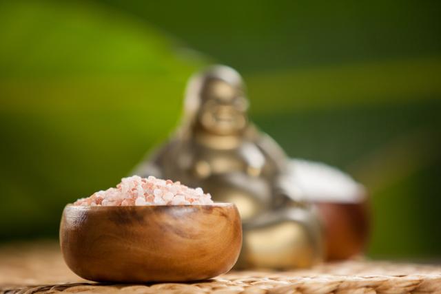 Laughing Buddha figurine and sea salt in wooden bowl on woven mat. Ideal for use in articles or advertisements related to relaxation, meditation, spa treatments, wellness, and spiritual decor. Perfect for promoting holistic and natural lifestyle products.